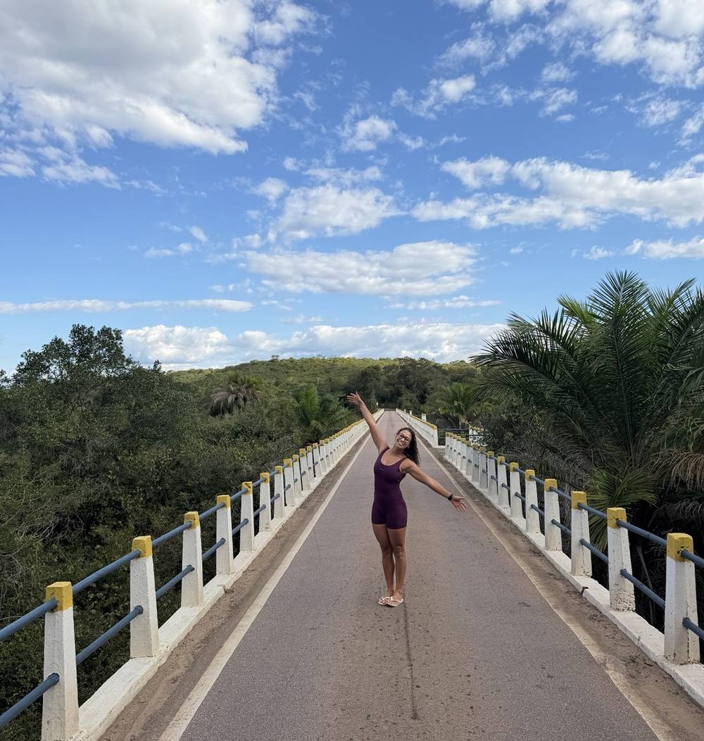 Michele de braços abertos no meio de uma ponte sob céu azul